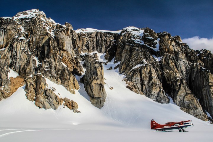 Red plane on snow near rocky, snowy mountains under a clear sky.