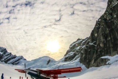 Small red plane on snowy mountain landscape under a cloudy sky.