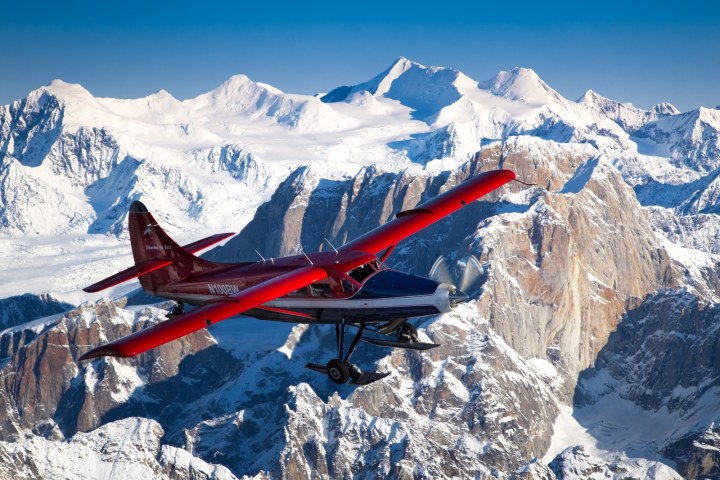 Red biplane flying over snow-covered mountains under a clear blue sky.