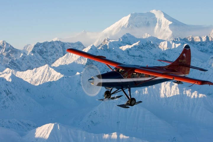 Red and blue small plane flying over snowy mountains with a clear sky.