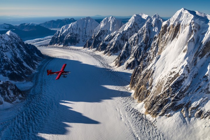 Red plane flying over snowy mountains and glacier under clear blue sky.