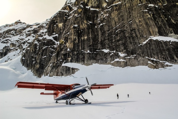 Small plane on snowy terrain near a steep rocky mountain, with three people walking nearby.